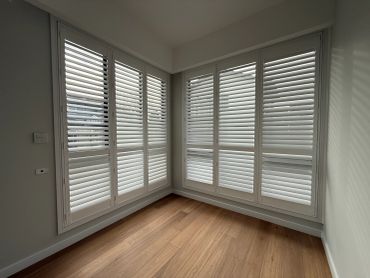 Empty corner room with white plantation shutters and wood floor.