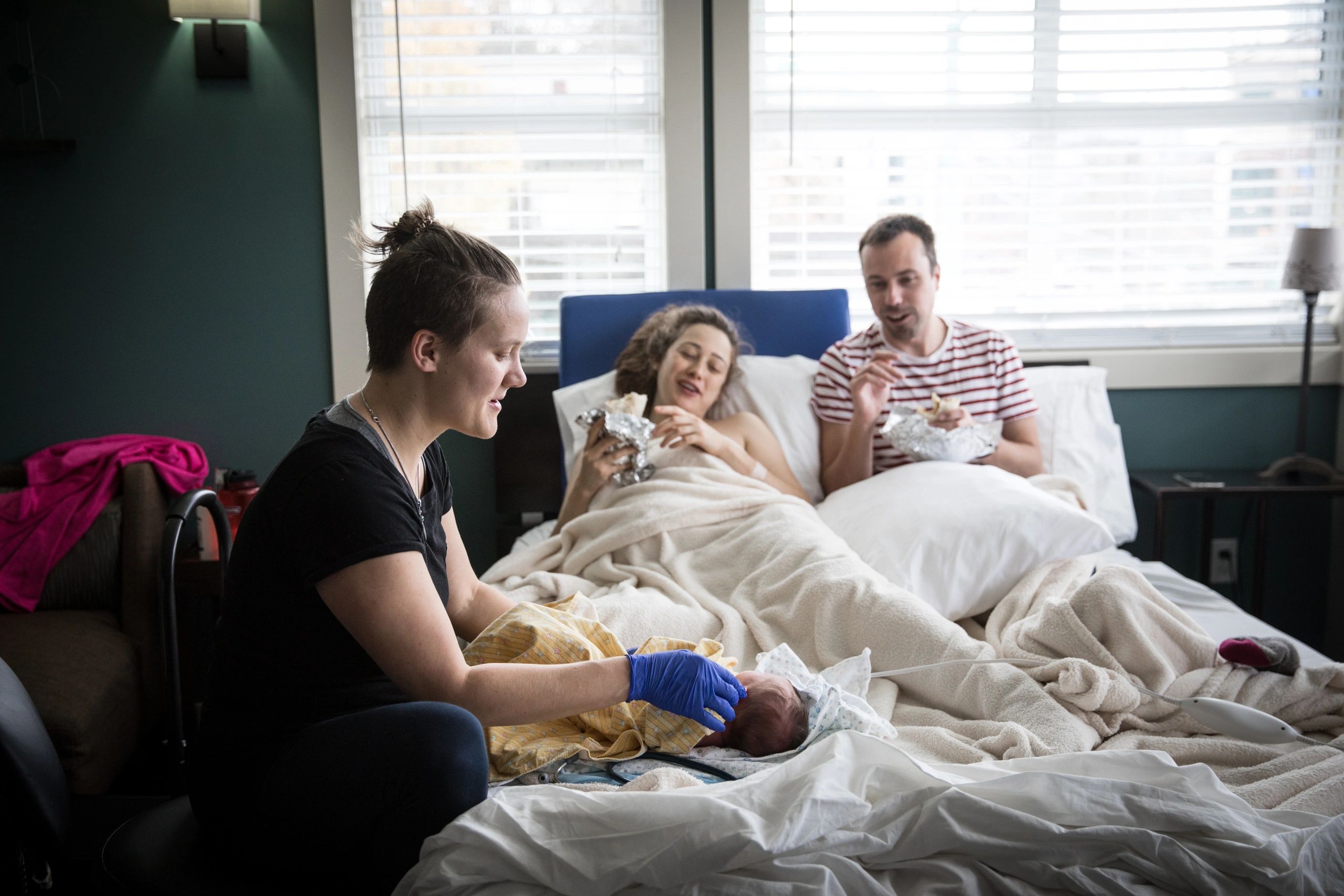 Wellspring Midwife Lizz examining a newborn while parents look on, smiling, eating burritos in bed. 