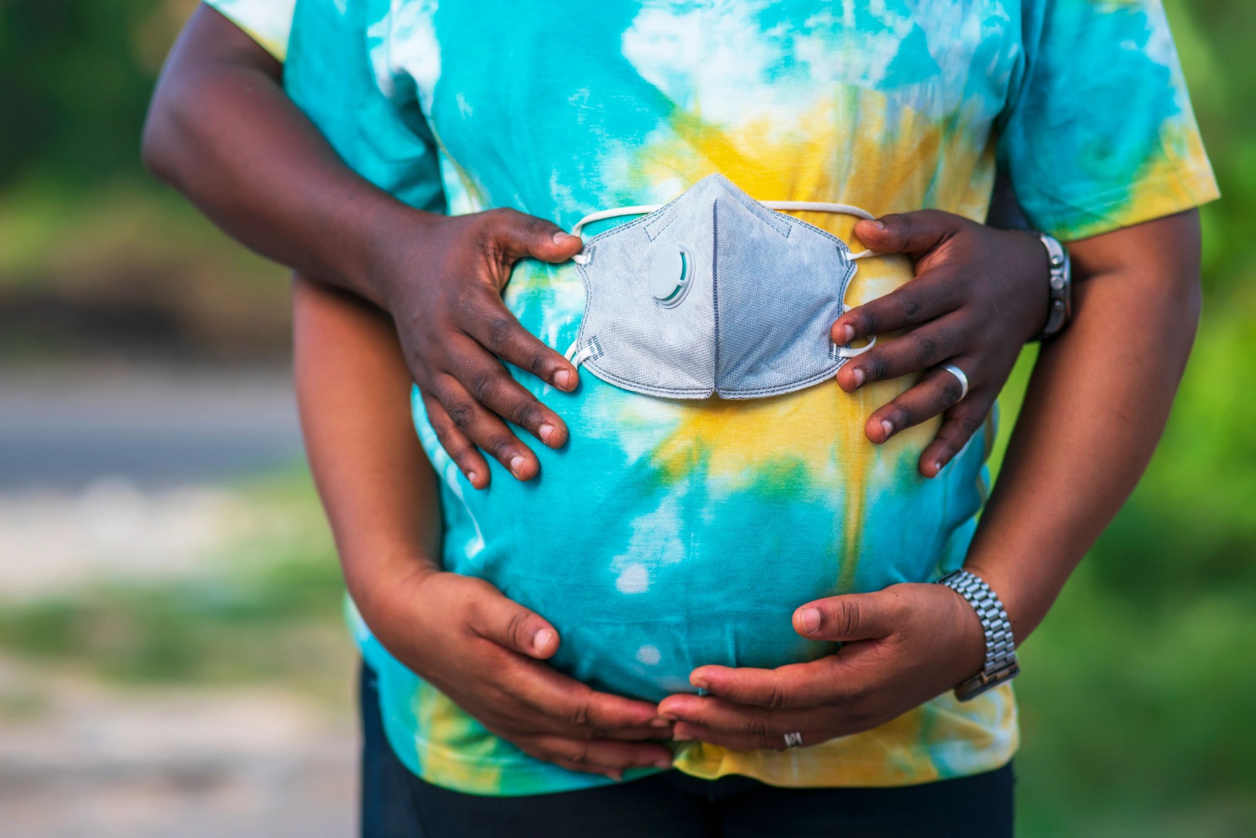 Photo of a Black person wearing a tie-dye shirt with a person behind holding a mask on their belly