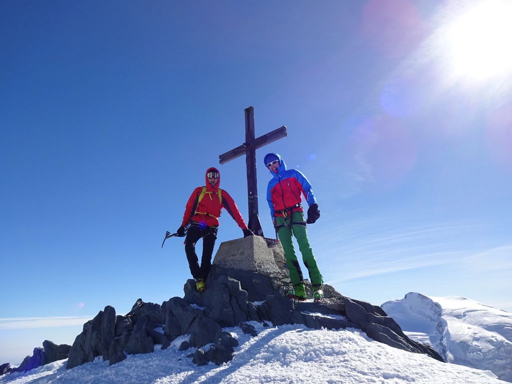 Two climbers stand next to a summit cross on a snowy mountain peak under a clear blue sky.