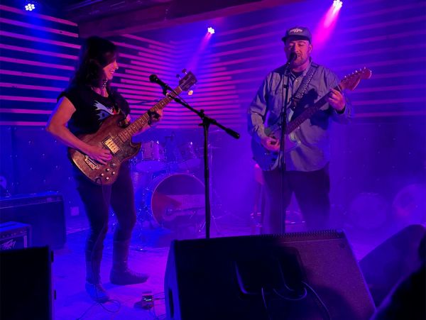 Two musicians playing guitars on a stage with purple lights.