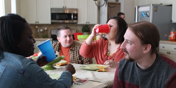 A group of people sitting around a table in a kitchen. They are talking, eating, and drinking.