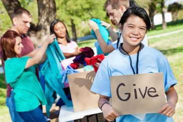 A young woman holds a sign saying 'Give' at a clothing donation event outdoors.