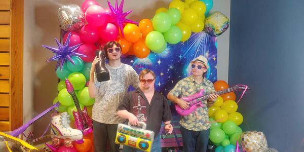 Three men with colorful props in a festive balloon backdrop.