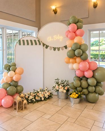 Baby shower backdrop with green, peach and coral balloon garlands flanking a round cream panel with 