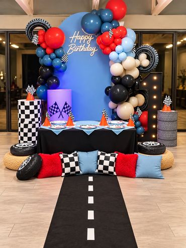 Race car themed birthday party setup with a red, blue and black balloon arch, checkered racing flags