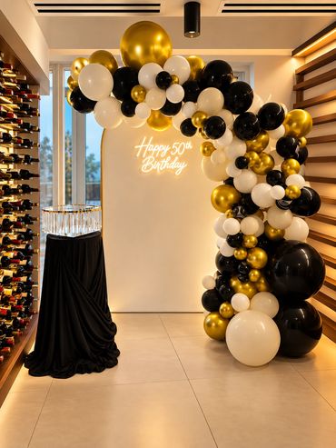 Elegant indoor event setup with a black, white and gold balloon garland arching over a black draped 