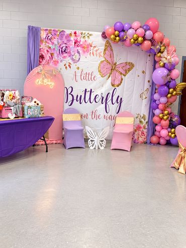Butterfly-themed baby shower celebration space with a pink, purple and gold balloon arch around an '
