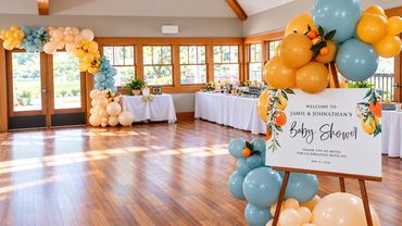 Spacious baby shower banquet hall with long dining tables dressed in white linens, blue and gold bal