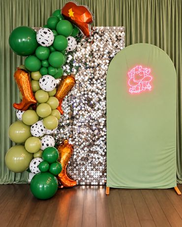 Dinosaur-themed birthday backdrop with a green balloon column featuring orange dinosaur foil balloon