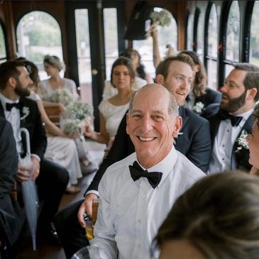 Smiling man in bow tie surrounded by wedding guests on a bus.