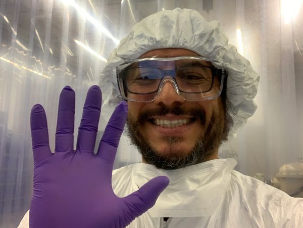 Smiling scientist in protective gear waves with a gloved hand.