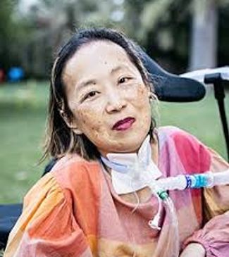 Alice, an Asian American woman with a tracheostomy, sits in a wheelchair wearing a colorful top.
