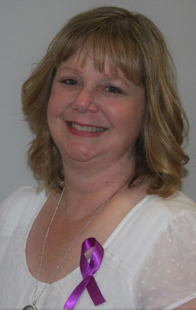 Smiling woman wearing a white blouse with a purple awareness ribbon.