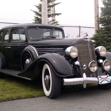 A classic black vintage car with whitewall tires parked near a fence and trees.