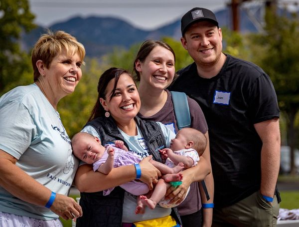 Four adults smiling together outdoors, with two holding babies.