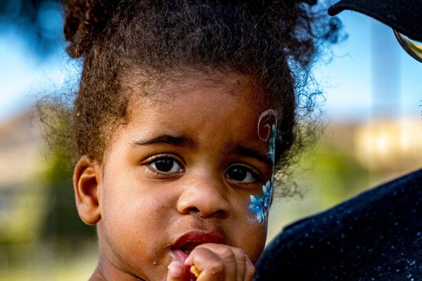 A young girl with face paint eating, held by a man wearing glasses and a hat.