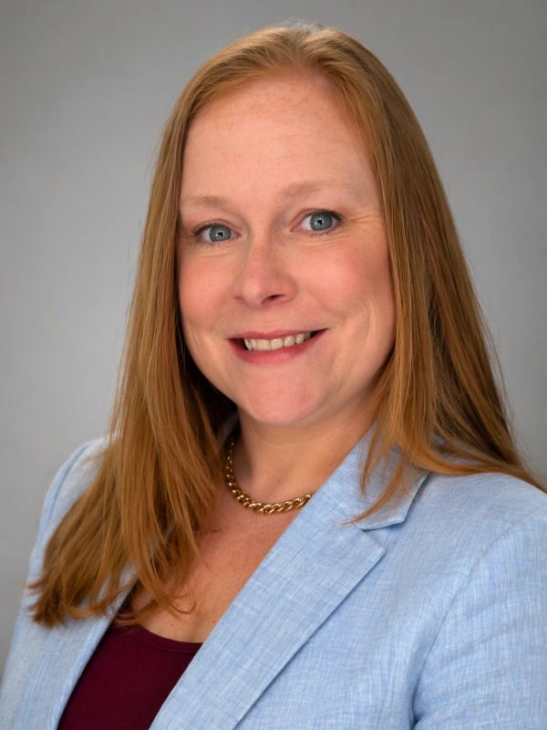 Professional woman with red hair wearing a light blue blazer and gold necklace.