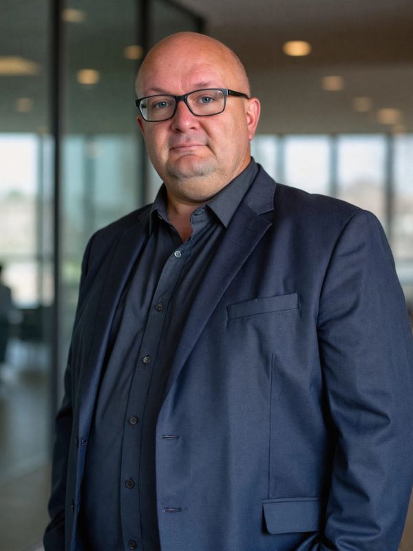 A man in a dark suit and glasses stands confidently indoors.