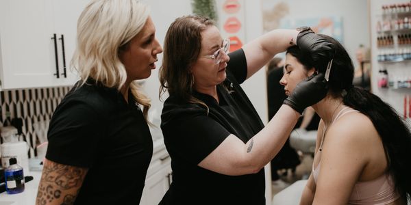 Two women training on ear piercing with a third woman as the model in a studio.