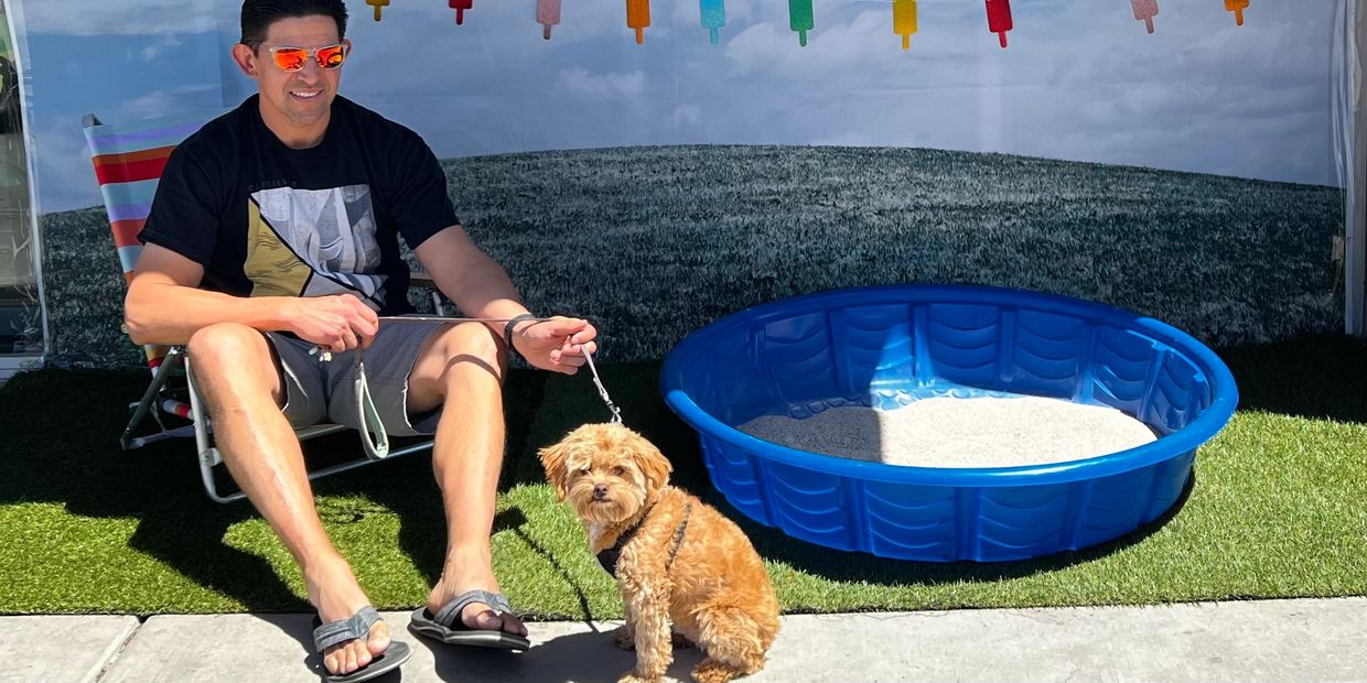 A man in sunglasses sits with a small dog near a blue kiddie pool outdoors.