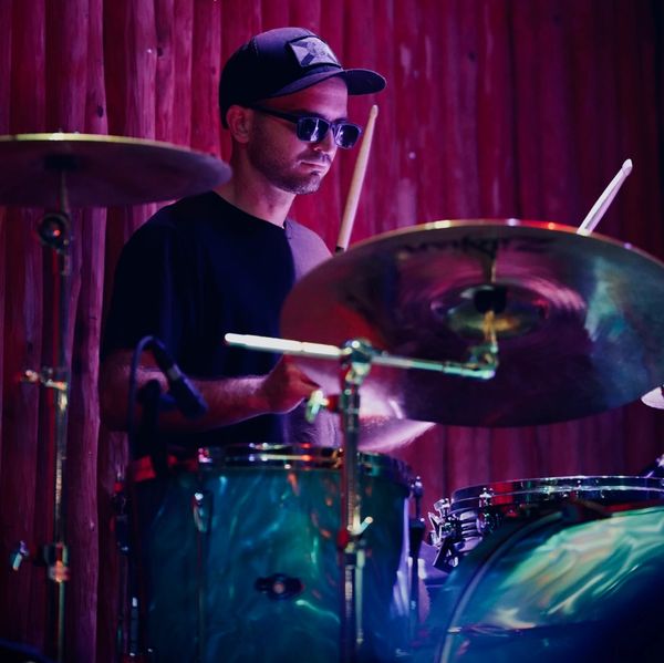 A drummer wearing sunglasses and a cap plays drums on stage.