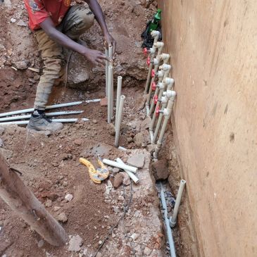 Worker installing PVC pipes in a dug trench beside a wall.