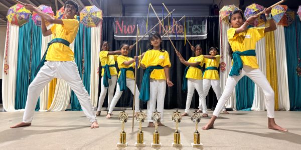 Children in yellow shirts performing a traditional stick dance on stage with trophies in front.
