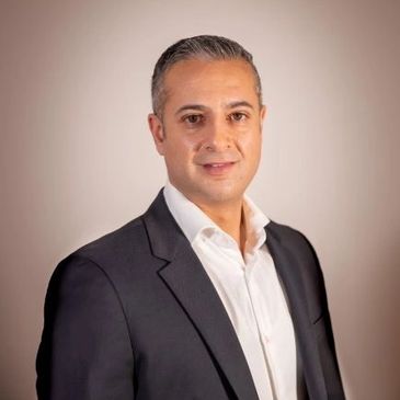 Professional man in a dark suit and white shirt posing against a neutral background.