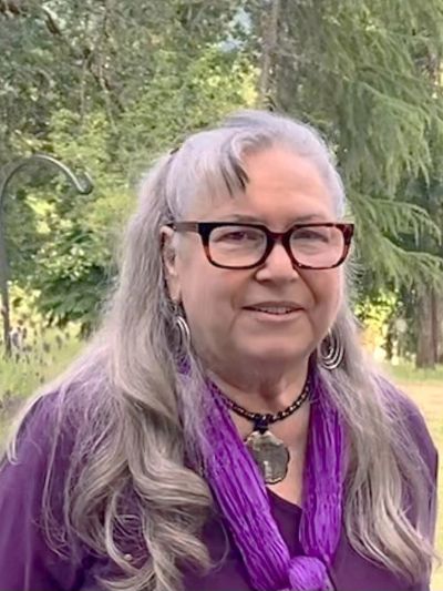 Elderly woman with long gray hair and glasses outdoors.