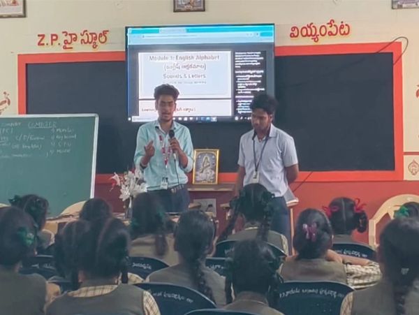 Two men teaching students in a classroom with a digital display.