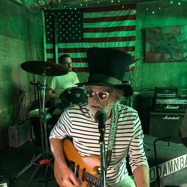 Musician in a striped shirt and top hat playing guitar with a drummer behind him.