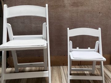 Two white folding chairs of different sizes placed on a wooden floor.