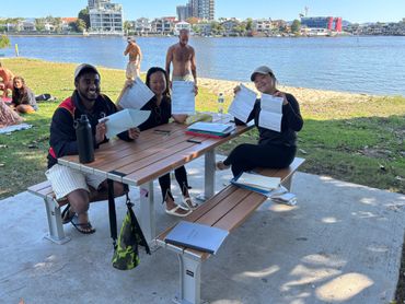 Three people happily holding documents at a picnic table by the water.