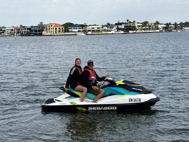 Two people enjoying a ride on a Sea-Doo jet ski on calm waters with houses in the background.