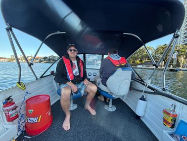 Two men wearing life jackets on a small boat with calm water and houses in the background.
