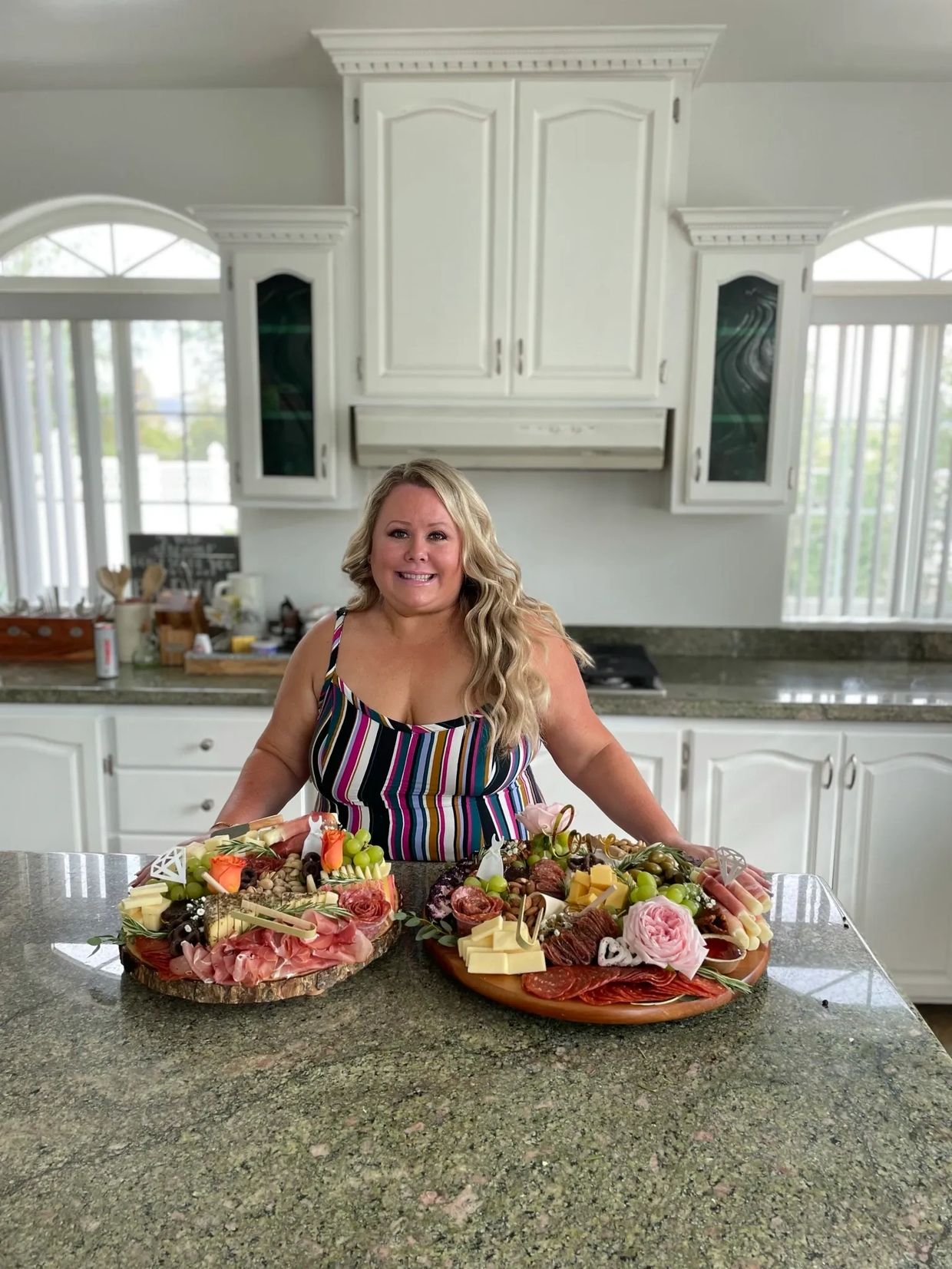 Woman smiling behind two large charcuterie boards in a bright kitchen.