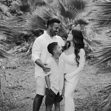 A happy family of three sharing a moment outdoors amidst palm leaves.