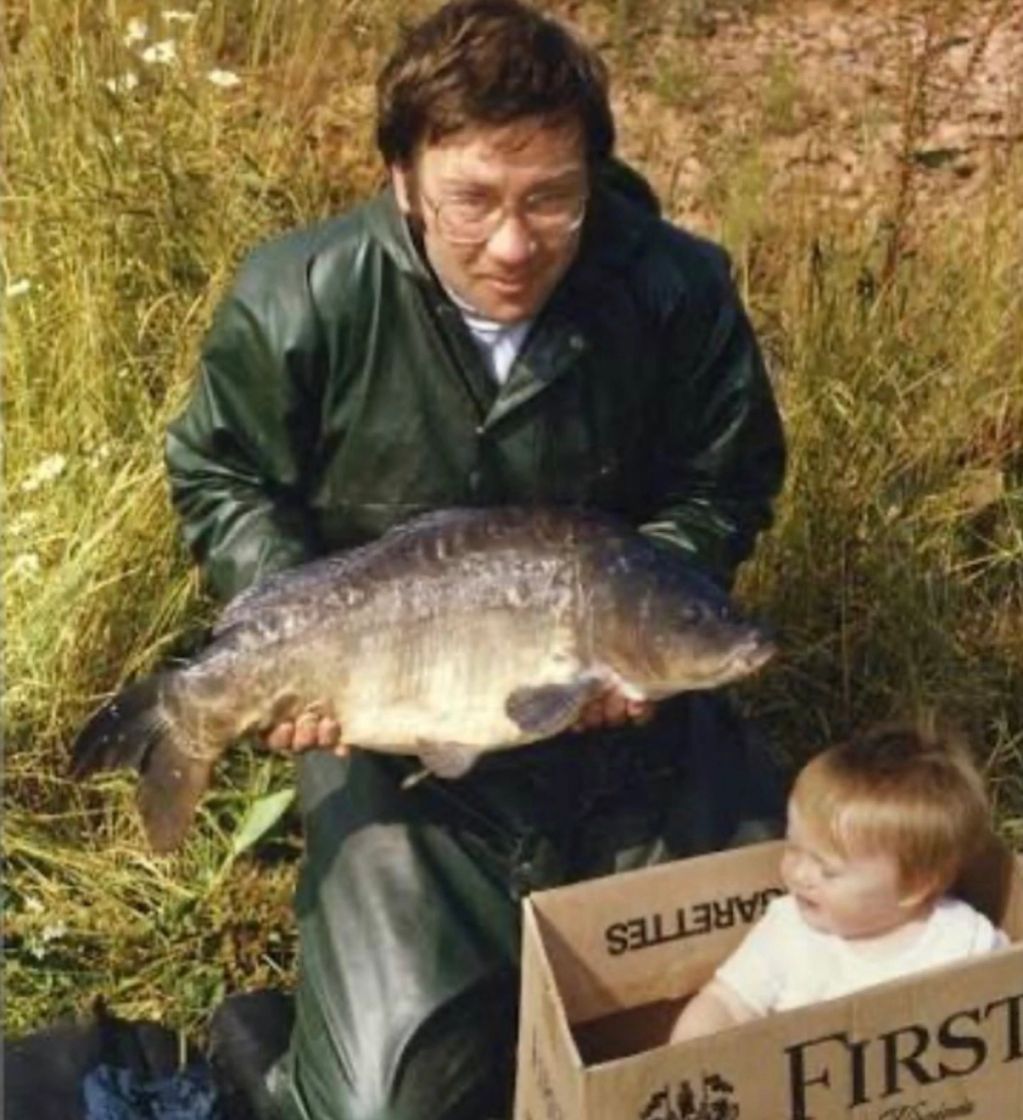 Man in raincoat holding a large fish, baby in a box next to him.