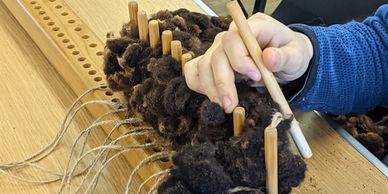 Person weaving dark wool fibers using wooden pegs on a loom.