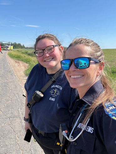 Two smiling EMS personnel posing outdoors on a sunny day.