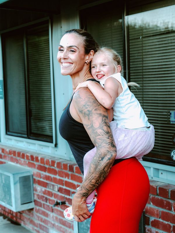Smiling woman with tattoos carrying a happy little girl on her back.