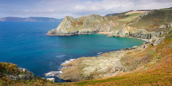 Tranquil coastal bay with rocky cliffs and clear blue water under a partly cloudy sky.