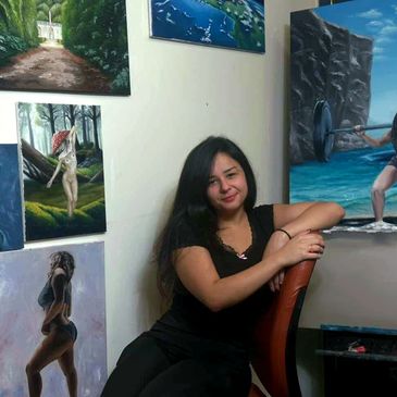A woman sits smiling in an art studio surrounded by paintings.