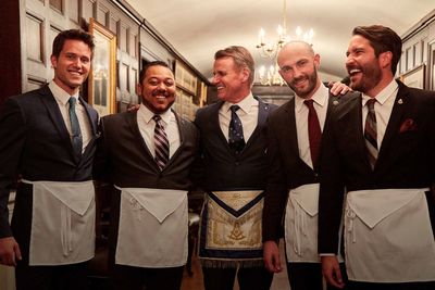 Five men in suits wearing Masonic aprons, smiling in a wood-paneled room.