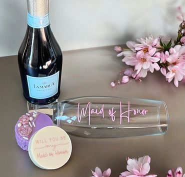 Maid of Honor gift set with prosecco, glass, and decorated cookies surrounded by pink flowers.