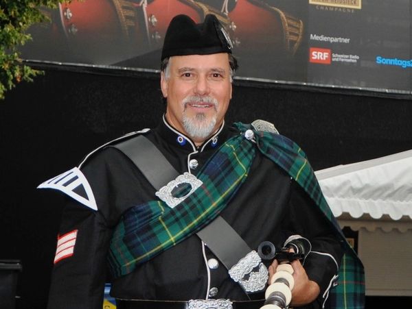 Man in traditional Scottish attire holding bagpipes and smiling.