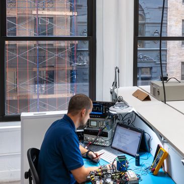 Technician working on electronic devices at a lab bench with windows showing an urban backdrop.