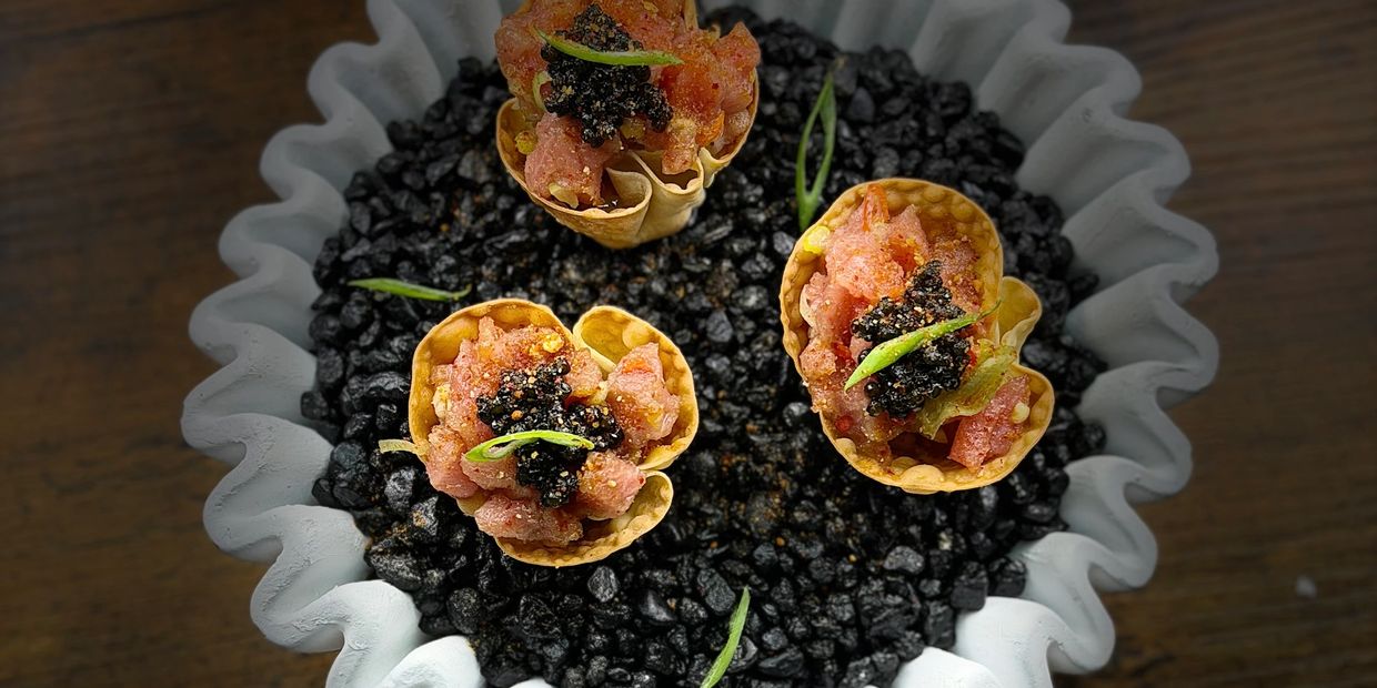 Three gourmet appetizers in small cones on black stones inside a ruffled white bowl.