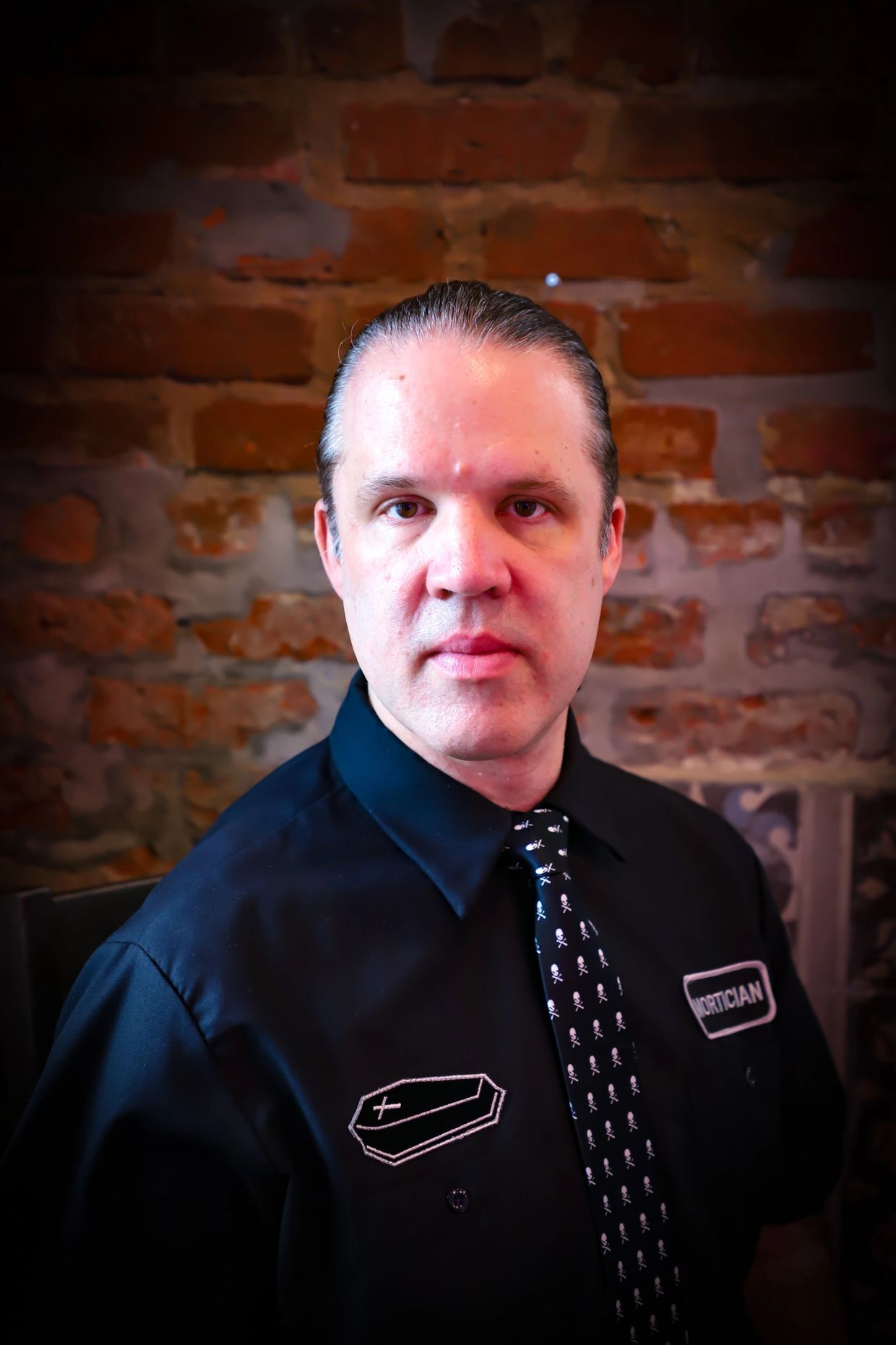 Man in a black shirt with mortician patches and skull tie posed against a brick wall.